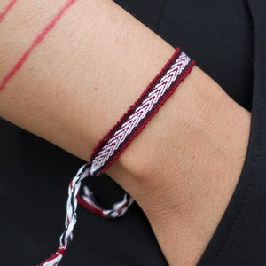 May include: A red and white woven bracelet with a braided design. The bracelet is tied with a knot and has a long tail.