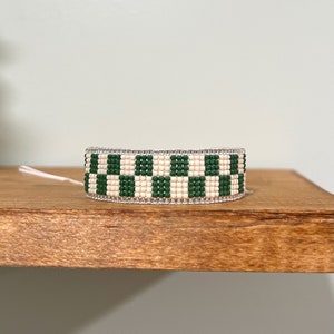 May include: A green and white checkered beaded bracelet. The bracelet is made with small, round beads and has a simple, modern design.