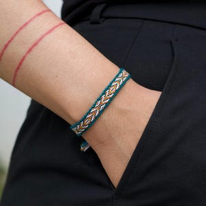 May include: A teal, white, and gold woven bracelet with a geometric pattern. The bracelet is worn on a wrist, with a black pants pocket in the background. The bracelet is approximately 0.5 inches wide.