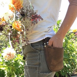 Op de afbeelding: Een persoon draagt een wit T-shirt en een bruine werkbroek met een bruine leren gereedschapstas die aan de riem is bevestigd. De tas heeft de tekst "Walter Moore" erop.