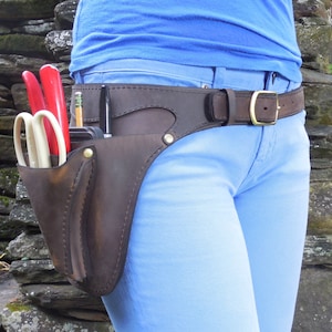 Op de afbeelding: Een bruine leren tuingereedschapsriem met een bruine leren holster voor scharen en een potloodhouder. De riem wordt gedragen over een blauwe jeans en een blauw shirt.