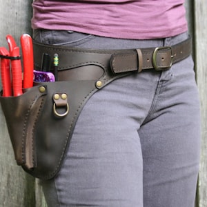 Könnte beinhalten: Ein brauner Leder-Werkzeuggürtel mit einer braunen Ledertasche an der Seite. Die Tasche ist mit roten und schwarzen Gartengeräten gefüllt. Der Gürtel wird über einer grauen Jeans getragen.