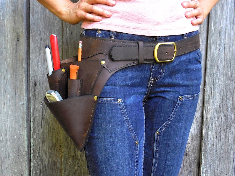May include: A brown leather tool belt with multiple pockets and a brass buckle. The belt is worn over blue jeans and a pink shirt.