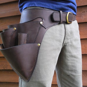 May include: A brown leather tool belt with a large pouch and multiple smaller pockets. The belt is worn over a pair of khaki pants.