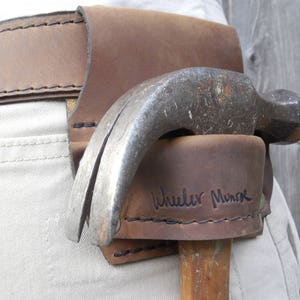 May include: A brown leather hammer holder with the text "Wheeler Monroe" stitched on it. A hammer is secured in the holder.