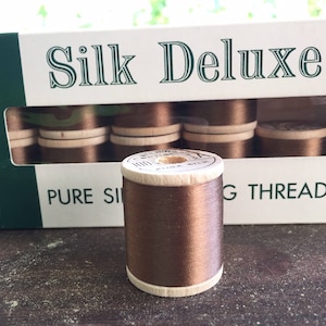 May include: A spool of brown silk sewing thread stands in front of a box of similar spools. The box is labeled "Silk Deluxe" and "Pure Silk Sewing Thread". The thread is wound on a wooden spool.