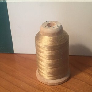 May include: A cylindrical spool of metallic gold thread wound around a light-colored wooden core. The top of the wooden spool features the printed black numbers 1228. The thread has a smooth, reflective finish and is tightly coiled around the center.