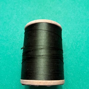 May include: A cylindrical spool of black sewing thread featuring a light-colored wooden core at the top and bottom ends. The black thread is wound tightly around the central section of the spool, with a single loose strand visible on the left side.