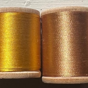 May include: Four spools of metallic thread in shades of red, gold, and bronze. The thread is wound around wooden spools with a light-colored rim. The colors range from a deep red to a bright gold and two shades of bronze.