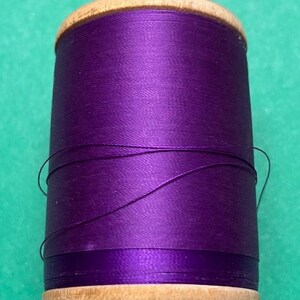 May include: A spool of vibrant purple sewing thread wrapped around a cylindrical wooden core. The thread has a smooth, slightly reflective finish, and a few loose strands are visible wrapped around the center of the spool.