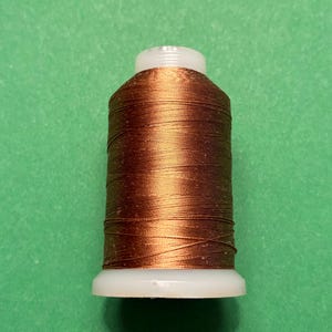 May include: A spool of metallic copper-colored embroidery thread wound tightly around a white plastic core. The thread has a smooth, reflective finish and is evenly distributed across the cylindrical body of the spool.
