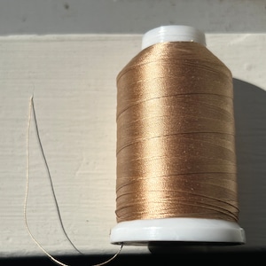 May include: A spool of metallic gold sewing thread wound around a white plastic cylinder. The thread has a smooth, reflective finish and a single loose strand extends from the base of the spool, showing the fine texture of the material.