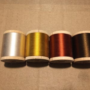 May include: Four wooden spools of thread arranged in a horizontal row. The spools feature metallic finishes in silver, gold, copper, and dark bronze. Each spool has a cylindrical shape with light-colored wooden ends and a smooth, shiny thread texture.
