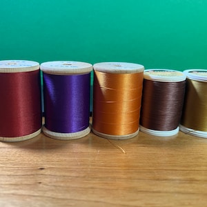 May include: Six spools of thread in various colors, including red, purple, orange, brown, and gold. The spools are made of wood and have a white label with text.