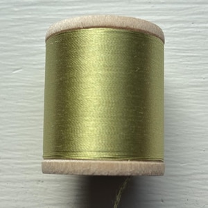 May include: A spool of olive green sewing thread wrapped around a cylindrical wooden core. The thread has a smooth, slightly lustrous finish, and the wooden spool features natural light-colored grain ends at the top and bottom of the cylinder.