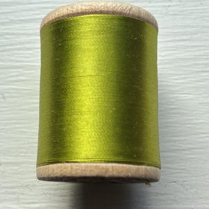 May include: A cylindrical wooden spool wrapped with vibrant lime green thread. The spool features natural light-colored wood ends with a smooth finish, while the central section is tightly wound with a continuous length of shiny, smooth green thread.