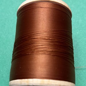 May include: A spool of dark brown sewing thread wrapped around a cylindrical wooden core. The thread has a smooth, slightly reflective finish and is wound tightly in horizontal layers from the top to the bottom of the light-colored wooden spool.