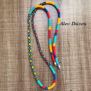 May include: A colourful beaded necklace with a toggle clasp. The necklace features a pattern of yellow, red, turquoise, and blue beads. The image includes the text "PDF TUTORIAL" and "Alev's Tutorials".