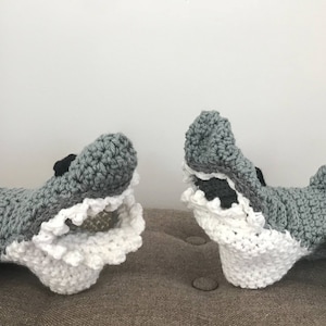May include: Two crocheted gray shark puppets with white frills around their mouths. The sharks have black eyes and are lying on a gray surface.