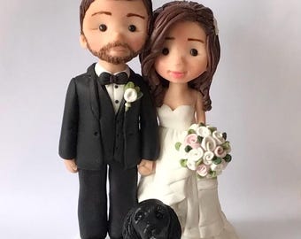 May include: A custom wedding cake topper featuring a bride and groom with a black dog. The bride is wearing a white dress and the groom is wearing a black suit. The dog is sitting in front of the couple.