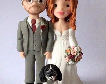 May include: A wedding cake topper featuring a bride and groom with a black and white dog. The bride is wearing a white dress and has red hair. The groom is wearing a gray suit and a red tie. The dog is sitting in front of the couple.