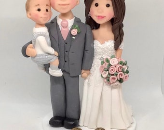 May include: A wedding cake topper featuring a bride and groom with a small child. The bride is wearing a white dress with a lace bodice and a bouquet of pink roses. The groom is wearing a gray suit with a pink tie. The child is wearing a white and gray outfit. There are two small brown turtles on the base of the topper.