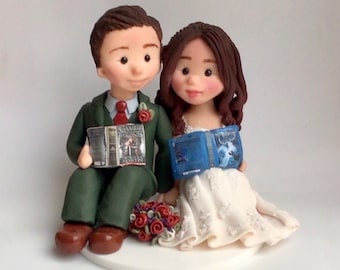 May include: A wedding cake topper featuring a bride and groom sitting side-by-side. The groom is wearing a green suit and holding a book titled "Game of Thrones". The bride is wearing a white dress and holding a blue book. They are both smiling.