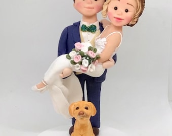 May include: A cake topper featuring a figurine of a person in a navy blue suit holding a person in a white wedding dress with a bouquet of pink and white flowers. A small yellow dog figurine sits at their feet on a round white base.