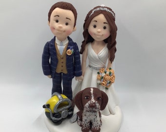 May include: A wedding cake topper featuring a bride and groom in a blue suit and white dress, holding a bouquet of flowers. A brown and white dog is standing in front of them, and a yellow diving helmet is on the base.
