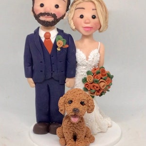 May include: Wedding cake topper featuring a bride and groom figurine with a small dog. The groom wears a navy blue suit with an orange tie and boutonniere. The bride wears a white wedding dress and holds a bouquet. The dog is brown.