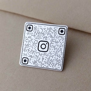 May include: A white metal pin with a black and white Instagram QR code design. The pin features a small Instagram logo in the center.
