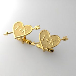 May include: A pair of gold-colored cufflinks. Each cufflink features a heart shape pierced by an arrow. The letters "I+K" are engraved on each heart. The cufflinks have a classic, elegant design.