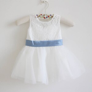 Pu&ograve; includere: Abito in tulle bianco con corpetto in pizzo e una cintura in raso blu chiaro.