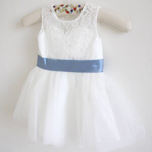 Pu&ograve; includere: Abito in tulle bianco con corpetto in pizzo e una cintura in raso blu chiaro.