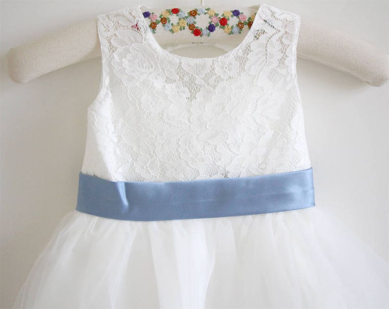Pu&ograve; includere: Abito bianco senza maniche in pizzo con un nastro di raso blu. L'abito ha una gonna di tulle ed &egrave; perfetto per una damigella d'onore.