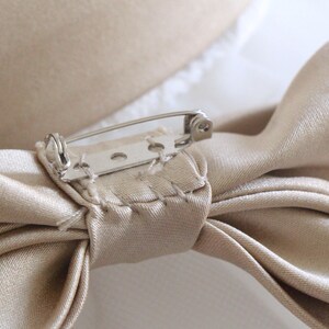May include: A beige satin bow with a silver safety pin attached. The bow is tied with a knot in the center and the ends are folded over.
