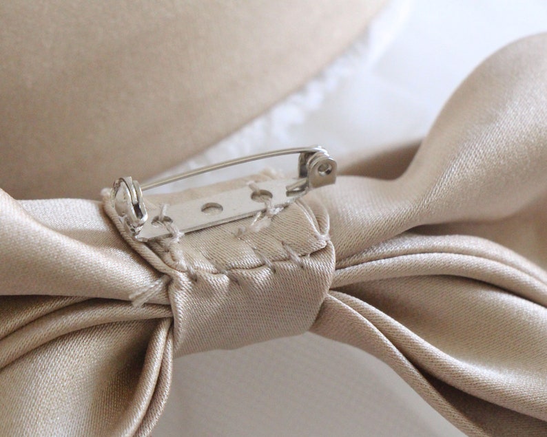 May include: A beige satin bow with a silver safety pin attached. The bow is tied with a knot in the center and the ends are folded over.