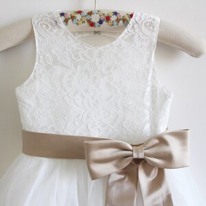 May include: White lace sleeveless dress with a tulle skirt and a wide, tan satin sash tied into a bow at the waist.