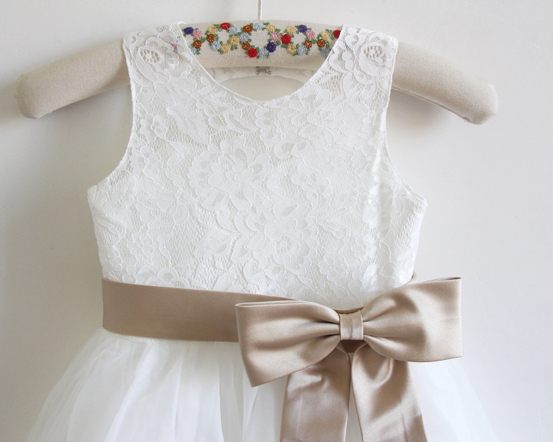 May include: White lace sleeveless dress with a tulle skirt and a wide, tan satin sash tied into a bow at the waist.