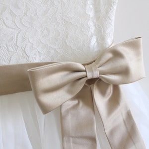 May include: A white tulle dress with a lace bodice and a beige satin sash tied into a bow.