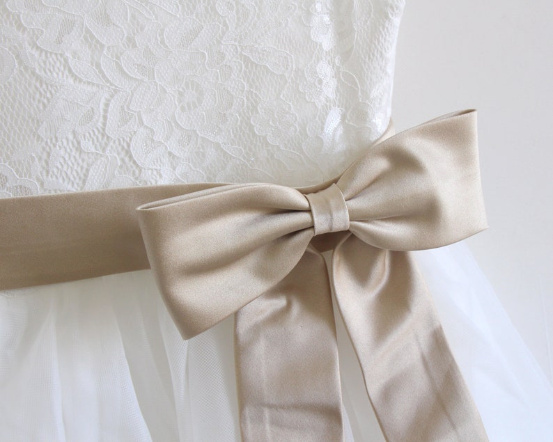 May include: A white tulle dress with a lace bodice and a beige satin sash tied into a bow.
