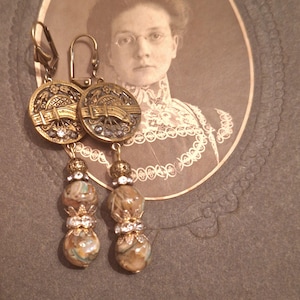 Puede incluir: Pendientes de estilo vintage con detalles metálicos dorados y cuentas colgantes. Los pendientes presentan adornados topes circulares con diseños intrincados y acentos brillantes. La parte inferior de cada pendiente tiene dos cuentas veteadas separadas por un elemento decorativo.