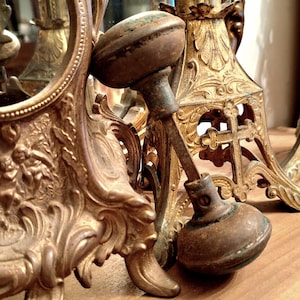 May include: An antique brass mirror frame and decorative candle holders. The mirror frame has an ornate design. The candle holders have a cross design. A vintage door knob is in the foreground. The items are in shades of gold and brown.