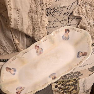 May include: An oval porcelain serving dish with a scalloped edge and a cream-colored finish. The dish features a repeating pattern of small, colorful portraits of women in period clothing, interspersed with delicate gold-toned floral filigree.