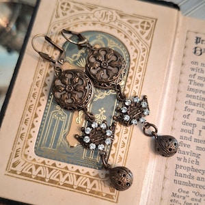 May include: A pair of bronze-toned dangle earrings featuring circular filigree medallions, small crown-shaped charms encrusted with clear rhinestones, and spherical filigree beads at the base, all suspended from secure lever-back ear wires.