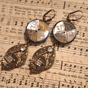 May include: A pair of dangle earrings featuring circular mother-of-pearl and gold-tone metal buttons at the top. Below, each earring has a bronze-tone filigree teardrop shape accented with a clear crystal and a small, textured circular metal charm.