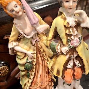 May include: Two porcelain figurines of a man and woman in 18th-century attire. The woman holds a pink parasol, and the man holds flowers. Both wear yellow coats, and the figurines have detailed facial features.