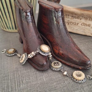 May include: Two antique wooden boot forms with a silver bracelet featuring decorative circular charms. The boots are dark brown and have a vintage appearance. The bracelet has a delicate design with cream-colored accents.