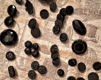 May include: A selection of black buttons in diverse shapes and sizes. The buttons include round, oval, and faceted designs, some with detailed patterns. The buttons are arranged on a vintage newspaper-style background.