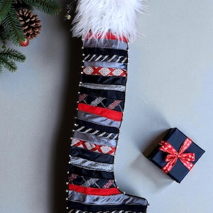 May include: A Christmas stocking made from repurposed neckties in black, red, and gray, with a white fluffy top. The stocking is decorated with small silver beads along the seams. A small black gift box with a red ribbon is next to the stocking.